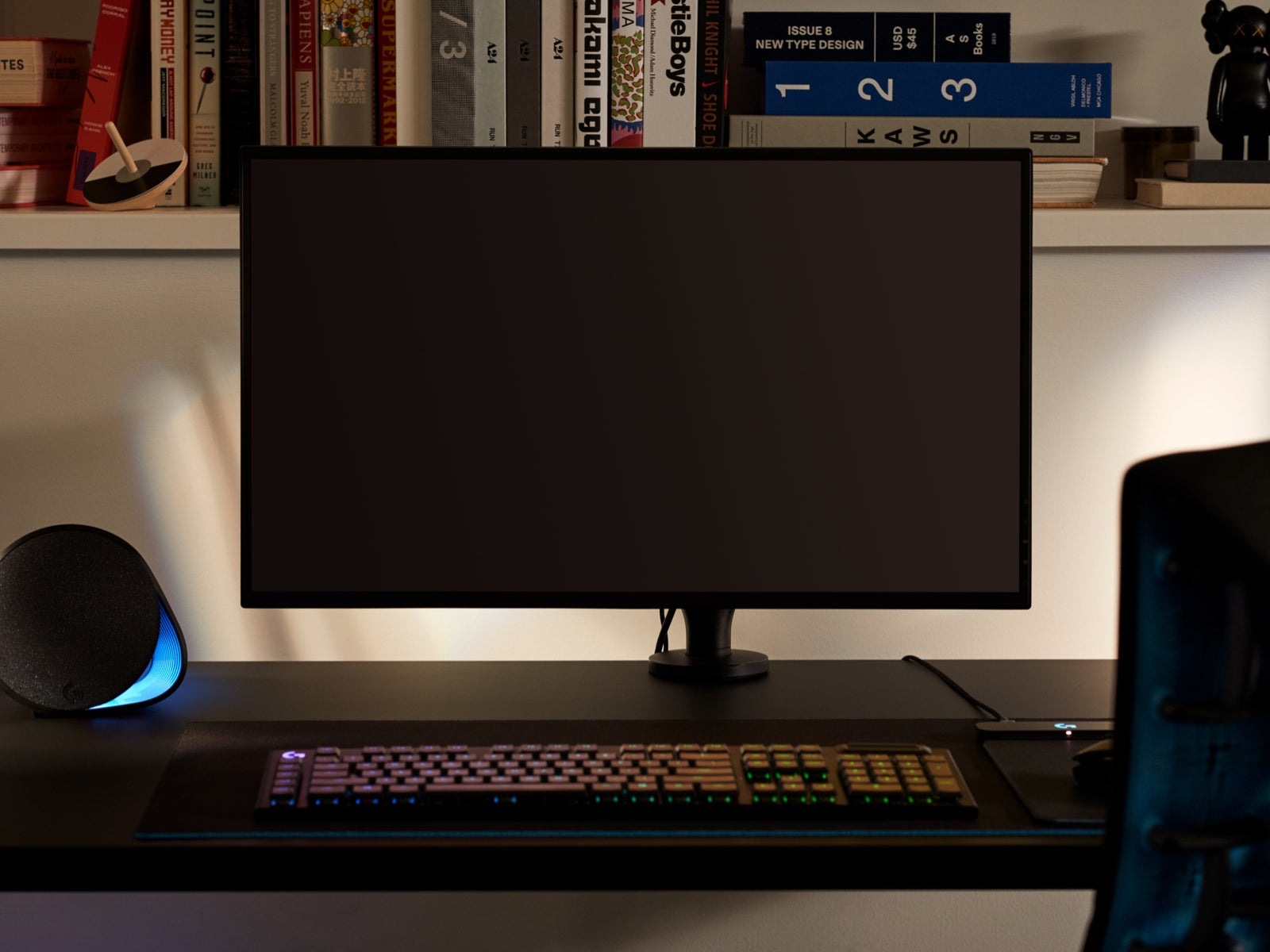 Un monitor grande, representando el hecho de que el brazo articulado para monitor Ollin puede acoplar pantallas de todos los tamaños, está unido al brazo en la parte superior de una mesa con un teclado en primer plano y artículos en una estantería de fondo.