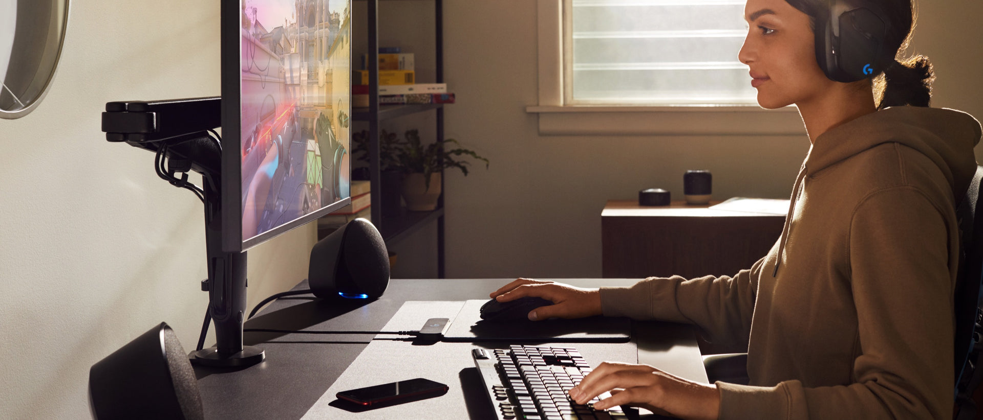 Una mujer con una sudadera con capucha y auriculares tiene una mano en el ratón y otra en el teclado mientras mira al monitor montado al brazo articulado para monitor Ollin.